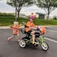 A smiling girl with cerebral palsy rides an adaptive tricycle while a younger girl helps her by pushing the handles.